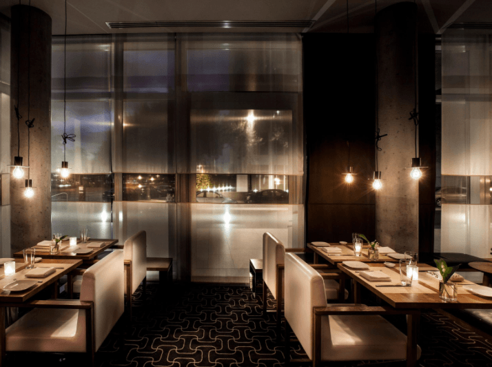 A modern, dimly lit restaurant interior with white cushioned chairs, wooden tables set for Pizza Suprema dining, pendant lights hanging above, and large frosted windows creating a cozy atmosphere.